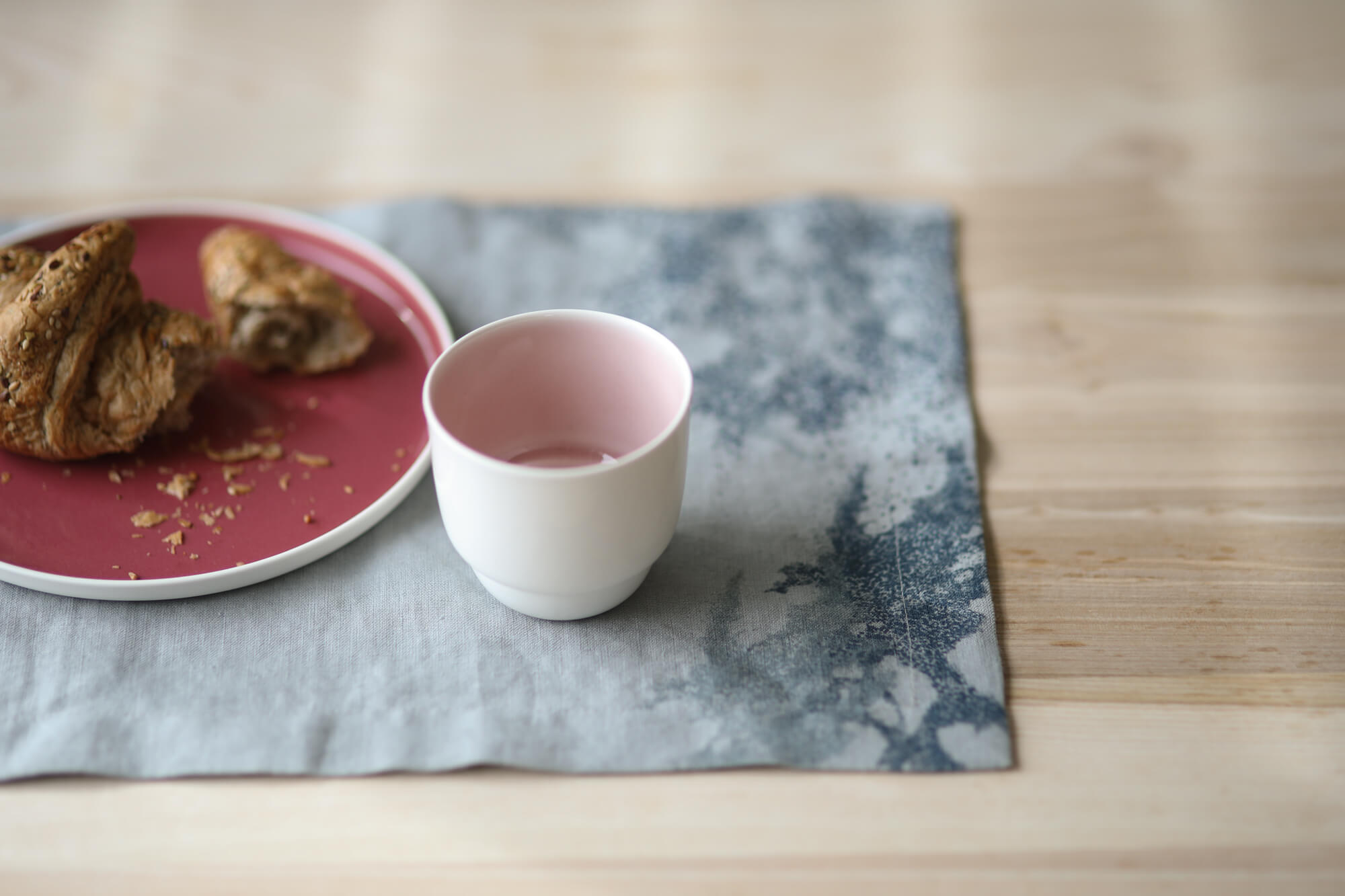 Na lnianej podkładce z nadrukiem Poppy stoi różowy, porcelanowy talerz z rogalem i porcelanowy kubek.