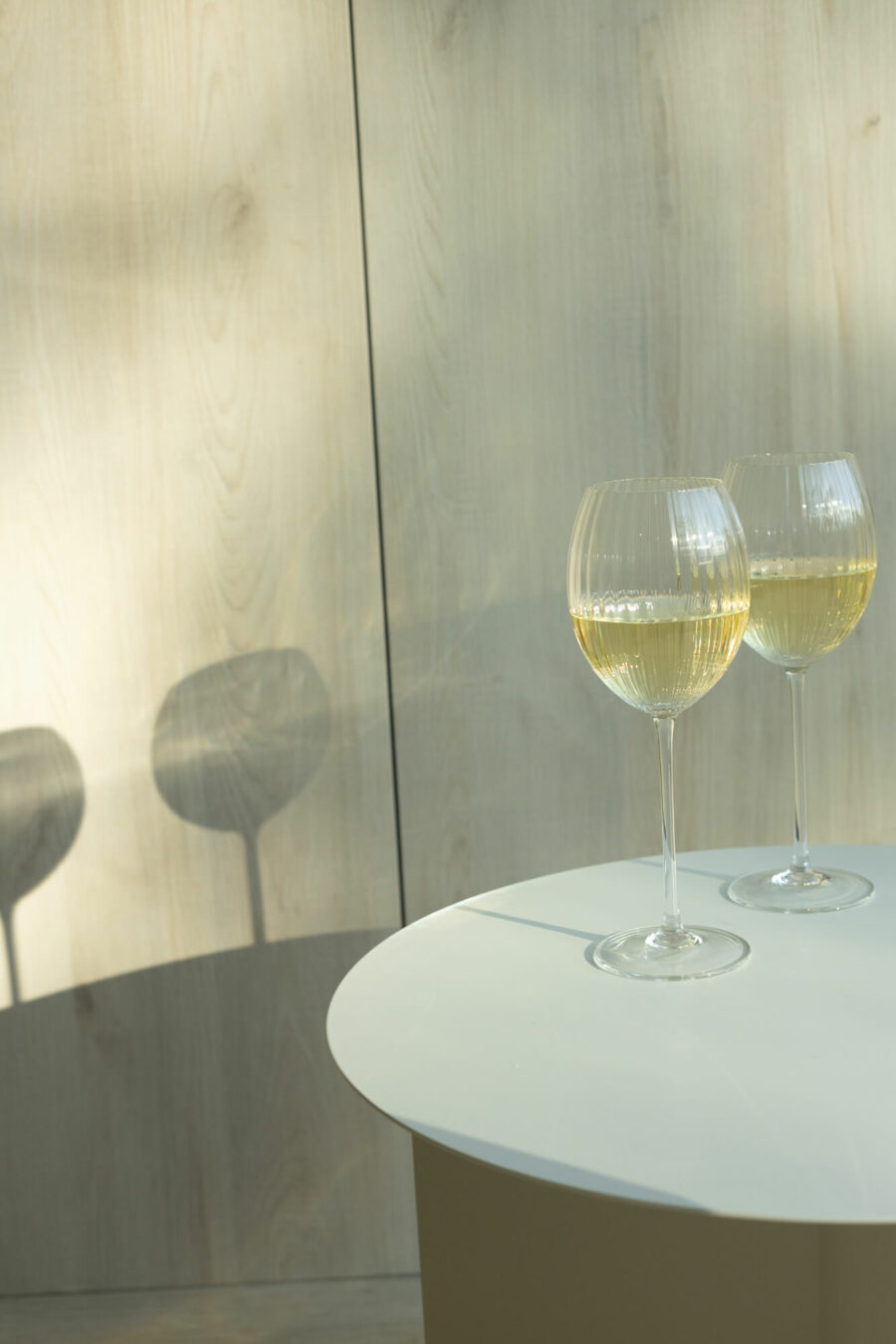 Na okrągłym stole stoją dwa kieliszki do białego wina. Na ścianie widać ich cień.