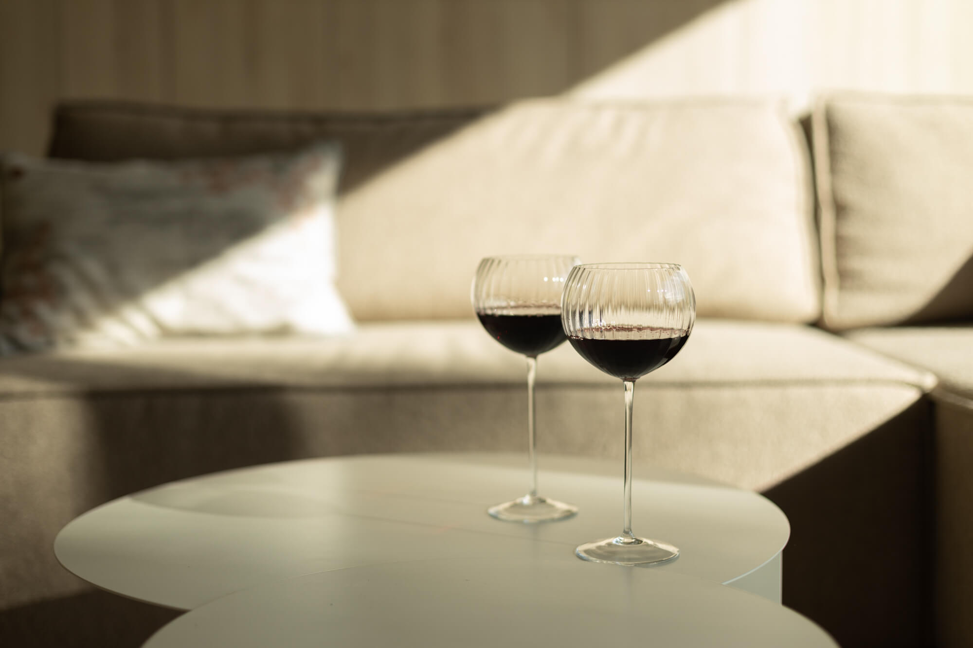 Na tle kanapy, okrągłym stole stoją dwa szklane, bezbarwne kieliszki do czerwonego wina.