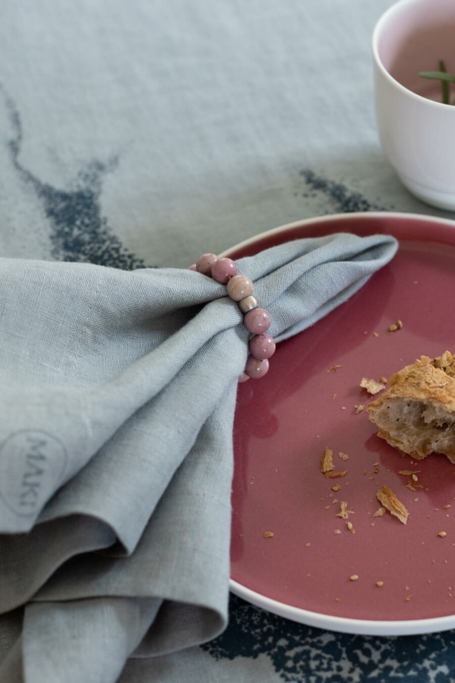 Kadr: od lewej: lniana serwetka obiadowa w obrączce z naturalnych kamieni różowego jaspisu, leży na różowym, porcelanowym talerzu, na nim kawałek bułki.
