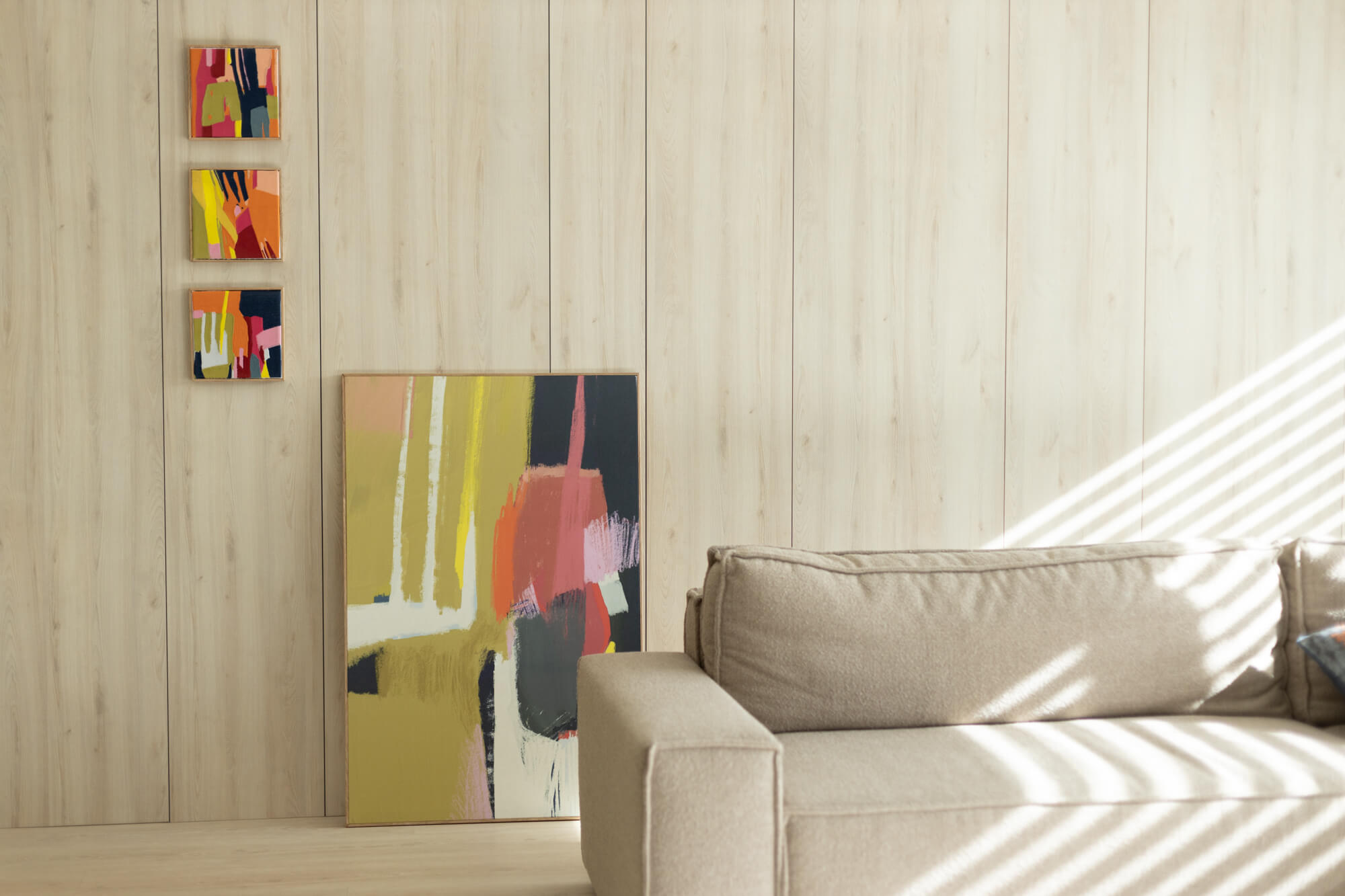 Wnętrze salonu. Na ścianie widzą trzy obrazki dekoracyjne w formie kwadratu, na podłodze stoi kolorowy obraz abstrakcyjny. Na pierwszym planie kanapa.