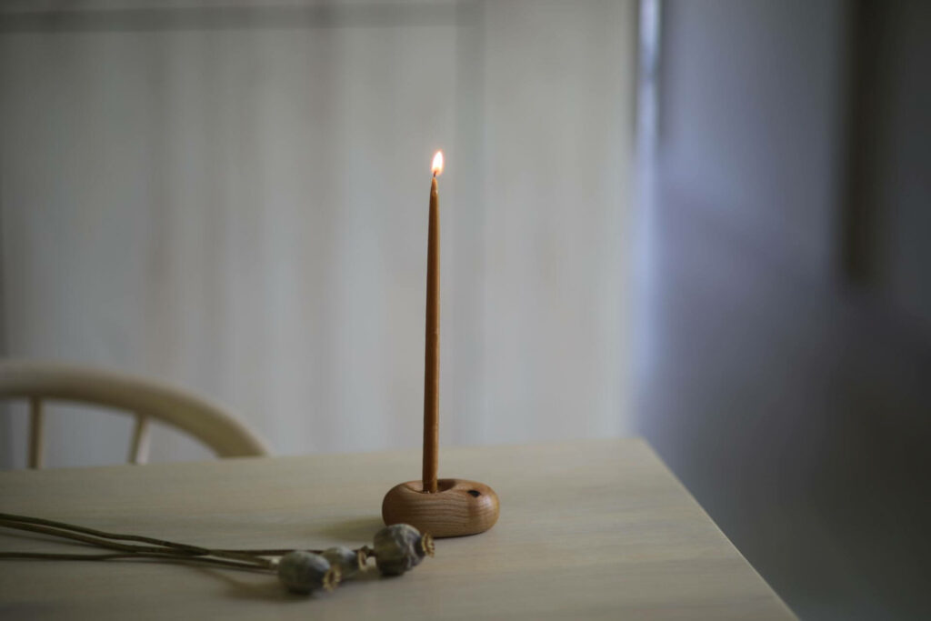 Na stole stoi świecznik z zapaloną wąską i długą świeczką. Przed nim leżą makówki.