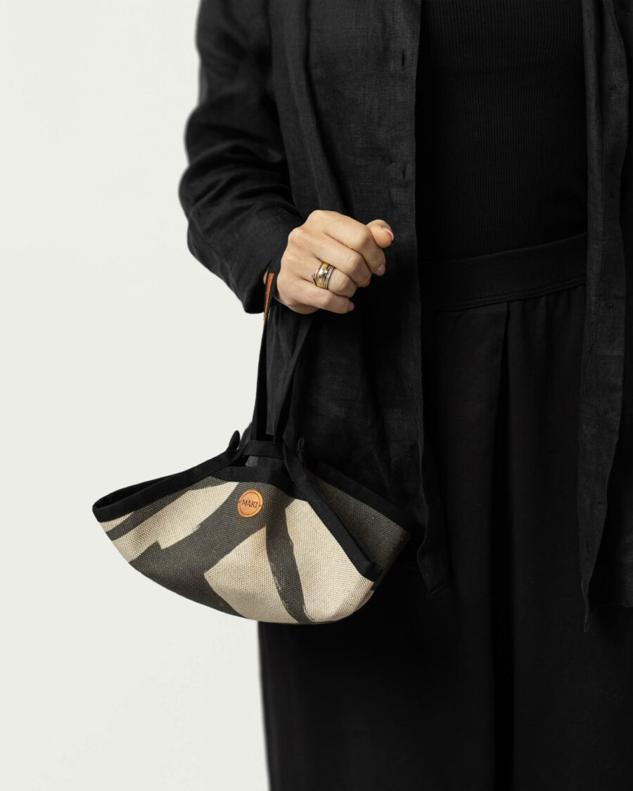 Stojąca, ubrana na czarno postać bez głowy trzymający w prawej ręce złożone lniane nosidło piknikowe. Małe nosidło dekorowane abstrakcyjnym geometrycznym printem.