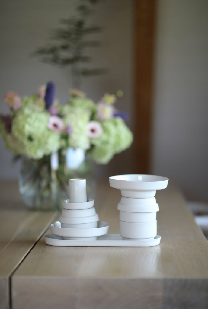Na drewnianym stole stoi komplet porcelanowy ponds. Naczynia ustawione są w wieżyczki. W tle bukiet zielonych i różowych kwiatów.